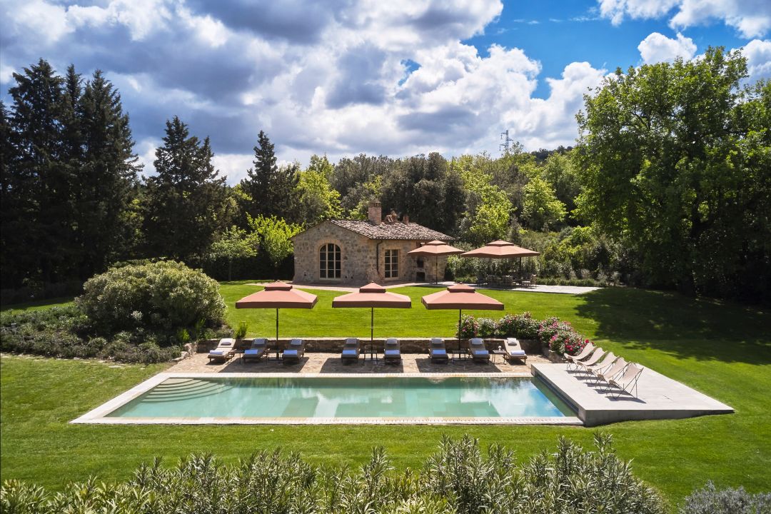 An outdoor pool surrounded by lounge chairs and umbrellas with a stone cottage behind